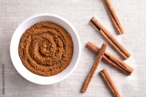Cinnamon powder, in a white bowl on linen fabric. Raw bark of Indonesian cinnamon, Cinnamomum burmannii, ground and whole. Used mainly as aromatic condiment and flavouring additive in a wide variety. photo