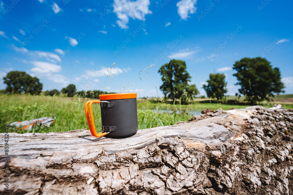 Camping aluminum mug stands on a log against the sky, tea utensils, metal glass, folding handles, silicone lip rim. Camping aluminum mug stands on a log against the sky, tea utensils, metal glass, folding handles, silicone lip rim.
