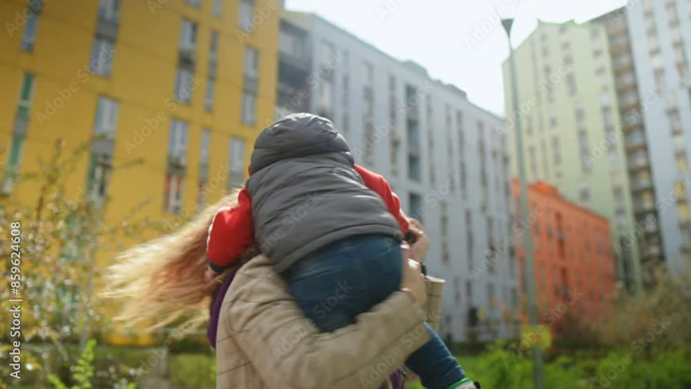 Slow motion. Close-up. A happy mother hugs her little son, holding him in her arms and spinning with him, standing in a sunny spring day in the courtyard of a modern residential complex Slow motion. Close-up. A happy mother hugs her little son, holding him in her arms and spinning with him, standing in a sunny spring day in the courtyard of a modern residential complex