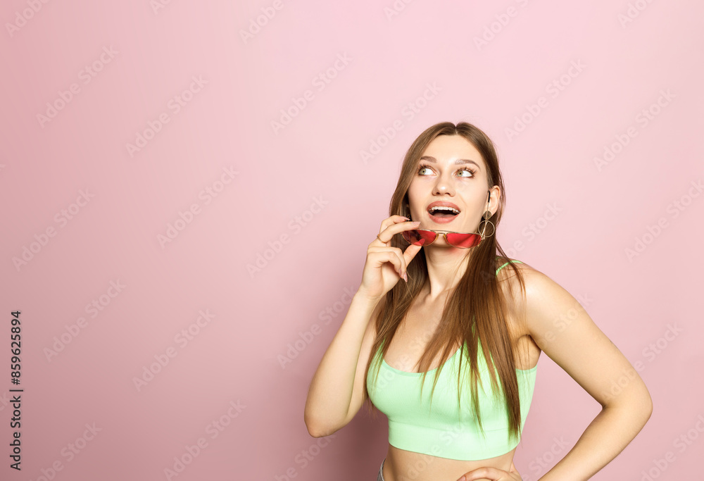 A woman is holding glasses and wearing a green top and shorts on a pink background. Beautiful figure and smile. Sexy look. Look from under glasses. A woman is holding glasses and wearing a green top and shorts on a pink background. Beautiful figure and smile. Sexy look. Look from under glasses.