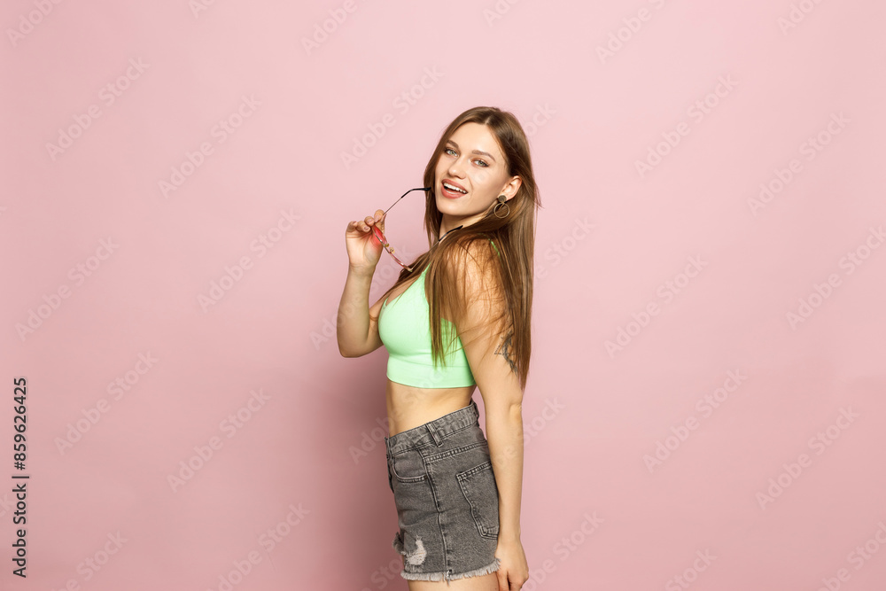 A woman is holding glasses and wearing a green top and shorts on a pink background. Beautiful figure and smile. Sexy look. Look from under glasses. A woman is holding glasses and wearing a green top and shorts on a pink background. Beautiful figure and smile. Sexy look. Look from under glasses.