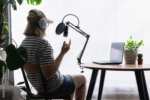 Podcast production concept. Young woman in big white headphones and baseball cap on white background with studio microphone, cup of tea and lap top with phrase On Air.