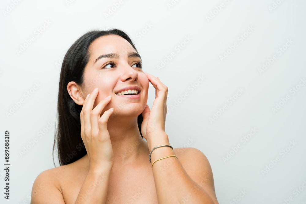Young latina woman is gently smiling and touching her radiant, clear skin in a bright studio setting Young latina woman is gently smiling and touching her radiant, clear skin in a bright studio setting