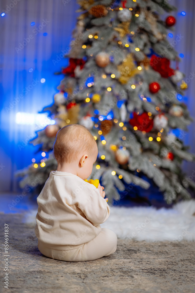 Baby Sitting by Christmas Tree. A baby in a white outfit sits on the floor, looking at a decorated Christmas tree. Baby Sitting by Christmas Tree. A baby in a white outfit sits on the floor, looking at a decorated Christmas tree.