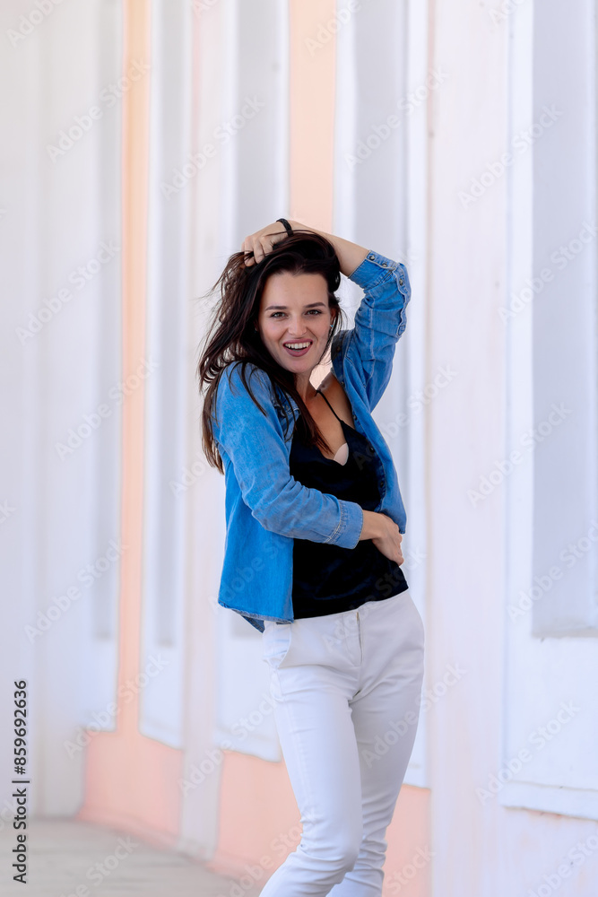 Fototapeta premium Woman in Blue Denim Jacket Smiles in Front of White Wall. A woman in a blue denim jacket and white pants smiles at the camera while standing in front of a white wall.