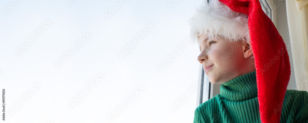 Children's dreams concept. Friendship. A lonely little boy in a Santa Claus hat is sad at the window waiting for gifts, looking out the window, the child is waiting for Santa Claus.