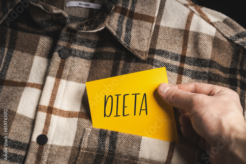 Yellow card with a handwritten inscription "Dieta", held in the hand against the background of a brown plaid shirt (selective focus), translation: Diet