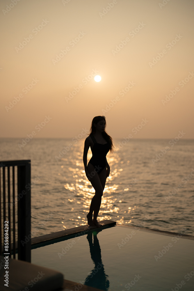 Fototapeta premium Woman in black swimsuit relaxing by infinity pool with ocean view during sunset.