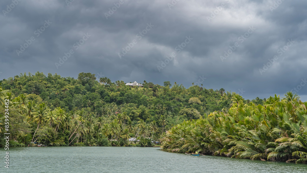 Fototapeta premium A beautiful turquoise river. There are impenetrable thickets of tropical vegetation and palm trees on the shores. A boat with a man in the riverbed. Roofs of houses among greenery. Clouds in the sky.