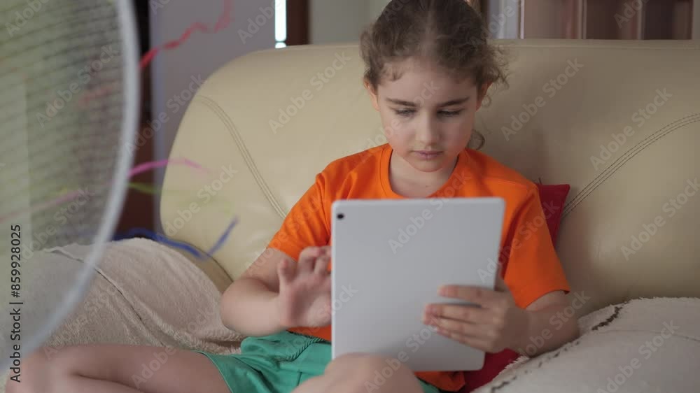 Girl Browsing Social Media on Tablet PC on Hot Heat Summer Day While Sitting on Sofa Near Fan. Child Plays Games on Tablet PC in Heat Weather Near Fan. Cool Wind From Electric Fan at Summer Heat. Girl Browsing Social Media on Tablet PC on Hot Heat Summer Day While Sitting on Sofa Near Fan. Child Plays Games on Tablet PC in Heat Weather Near Fan. Cool Wind From Electric Fan at Summer Heat.