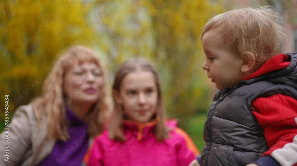 Slow motion. In the foreground, a young son stands thoughtfully in profile to the camera outdoors in a city park on a spring day. In the background, out of focus, the mother and teenage sister look at Slow motion. In the foreground, a young son stands thoughtfully in profile to the camera outdoors in a city park on a spring day. In the background, out of focus, the mother and teenage sister look at