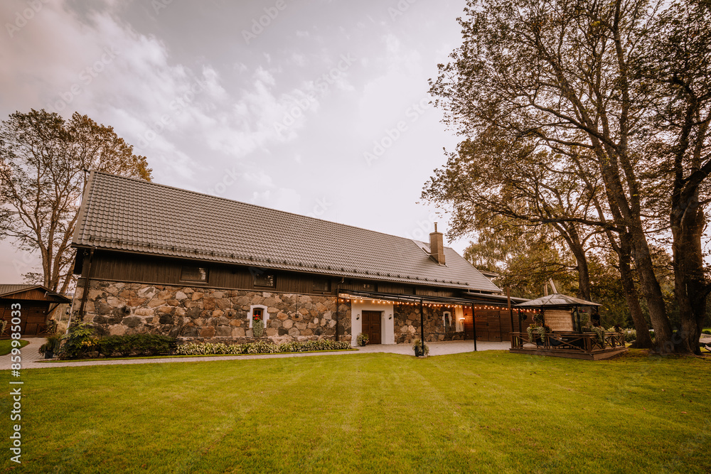 Blome, Latvia - September 11, 2023 - A rustic house with a stone and wood facade, surrounded by a lush green lawn and large trees, under a partly cloudy sky. Copy space. Blome, Latvia - September 11, 2023 - A rustic house with a stone and wood facade, surrounded by a lush green lawn and large trees, under a partly cloudy sky. Copy space.