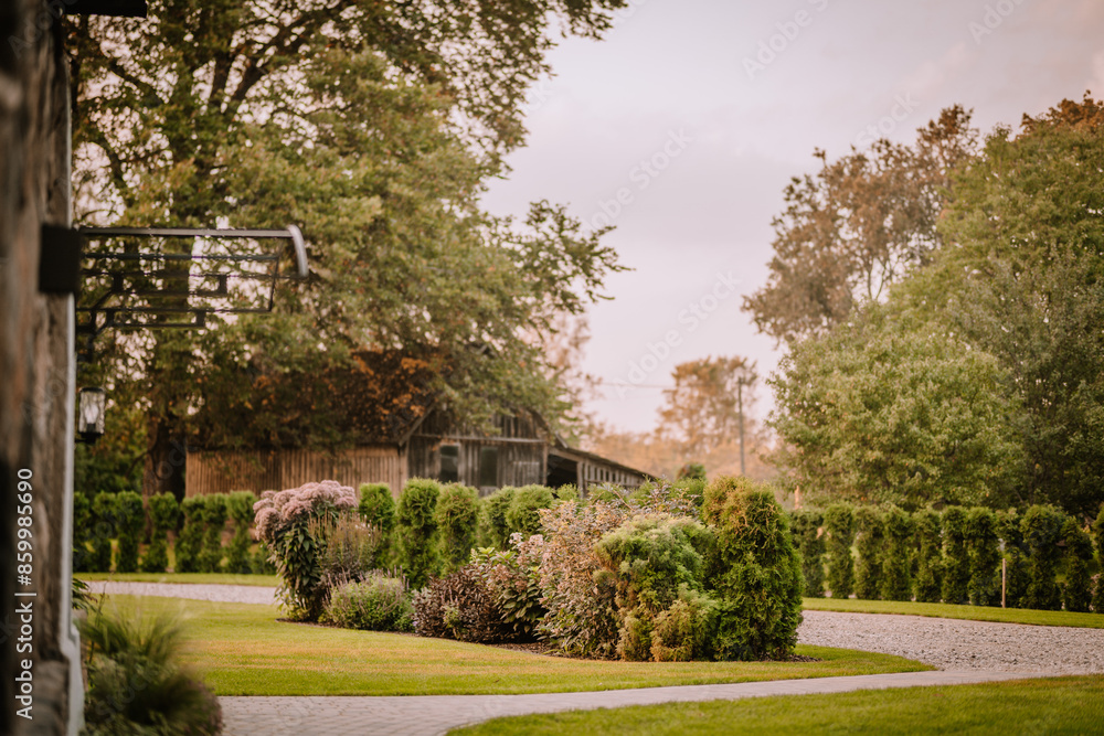 Blome, Latvia - September 11, 2023 - Beautiful garden with lush green bushes and trees, a gravel path, and a rustic wooden building in the background. Copy space... Blome, Latvia - September 11, 2023 - Beautiful garden with lush green bushes and trees, a gravel path, and a rustic wooden building in the background. Copy space...