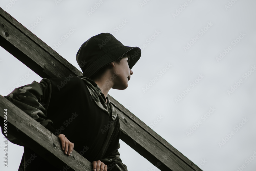 Young boy in a black hoodie and camo-patterned pants, wearing a blue hat, looks out from a wooden structure against a cloudy sky. Ample copy space. Young boy in a black hoodie and camo-patterned pants, wearing a blue hat, looks out from a wooden structure against a cloudy sky. Ample copy space.