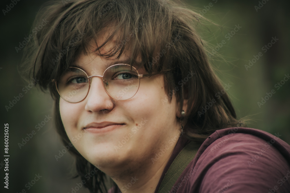 Close-up portrait of a teenager with glasses and a cross earring, wearing a maroon shirt, relaxing outdoors against a blurred green background. The teenager has a gentle smile. Close-up portrait of a teenager with glasses and a cross earring, wearing a maroon shirt, relaxing outdoors against a blurred green background. The teenager has a gentle smile.