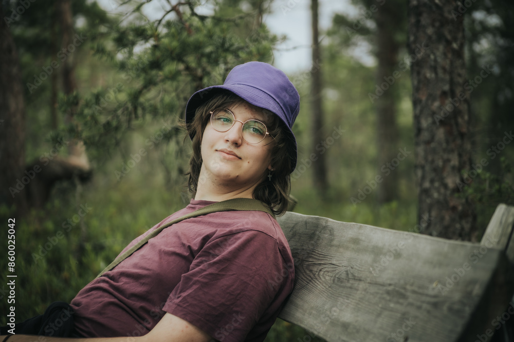 A teenager wearing a purple bucket hat and glasses leans against a wooden structure outdoors. They are dressed in a maroon shirt and have a relaxed expression. The background is blurred greenery. A teenager wearing a purple bucket hat and glasses leans against a wooden structure outdoors. They are dressed in a maroon shirt and have a relaxed expression. The background is blurred greenery.