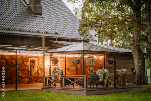 Blome, Latvia - September 11, 2023 - Cozy wooden gazebo with hanging plants and string lights, adjacent to a stone house, creating a warm and inviting atmosphere in the evening. Copy space... photo