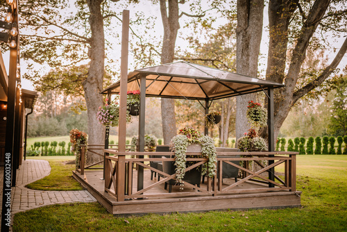 Blome, Latvia - September 11, 2023 - Charming wooden gazebo with hanging plants and outdoor furniture, surrounded by lush greenery and warm evening light, creating a cozy atmosphere. Copy space. photo