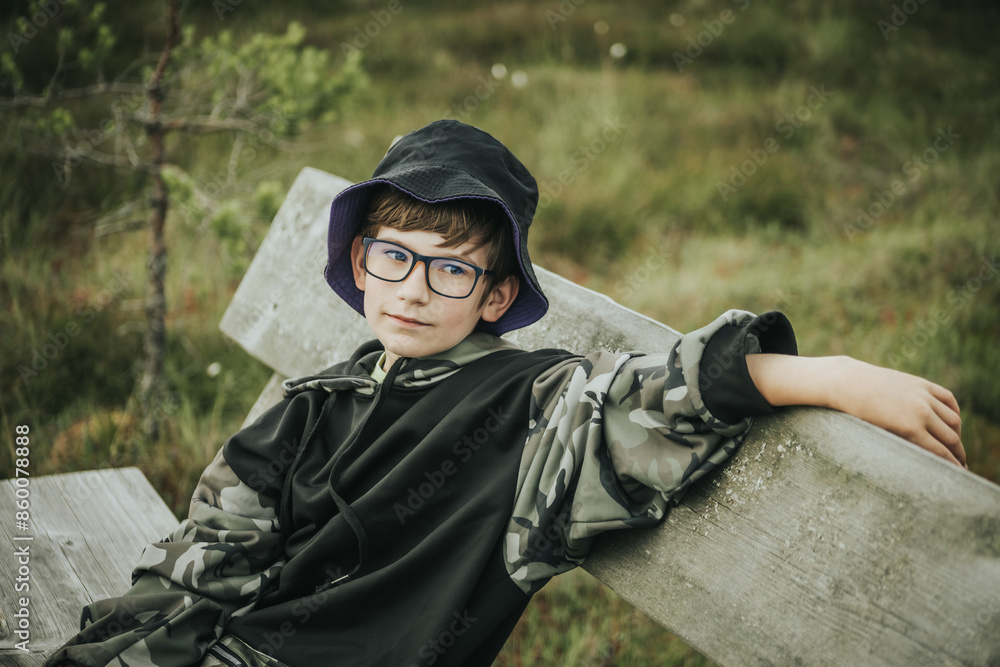 boy wearing glasses and a black bucket hat with a purple brim, sitting on a wooden bench outdoors. He wears a camouflage hoodie and has a playful expression on his face. boy wearing glasses and a black bucket hat with a purple brim, sitting on a wooden bench outdoors. He wears a camouflage hoodie and has a playful expression on his face.