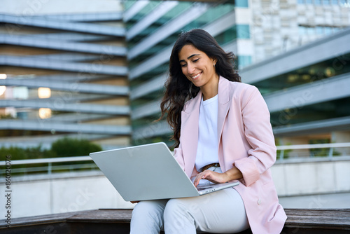 Smiling young middle eastern Israel businesswoman using online app on laptop pc. Indian or arabic woman freelancer in suit doing business analytics on computer, working remotely outdoors. Copy space