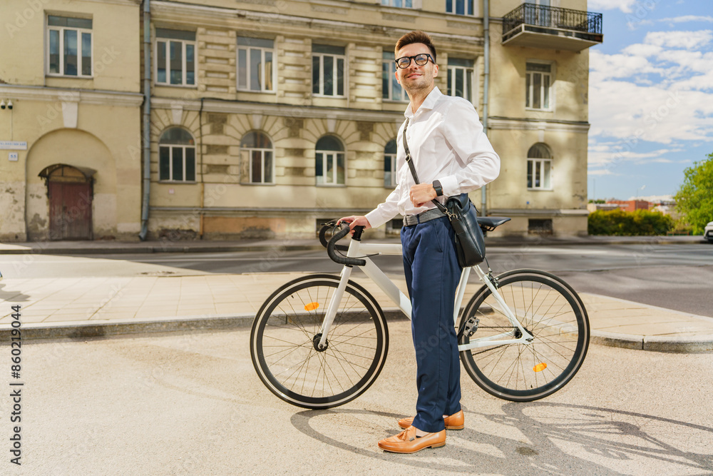 Obraz premium A young businessman, standing confidently with his bicycle and leather shoulder bag in an urban environment.