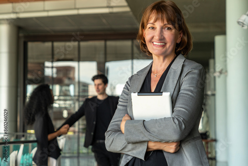 senior female executive in a suit holds a tablet with confidence and success. In the background, there are male and female employees talking together in the office corridor.