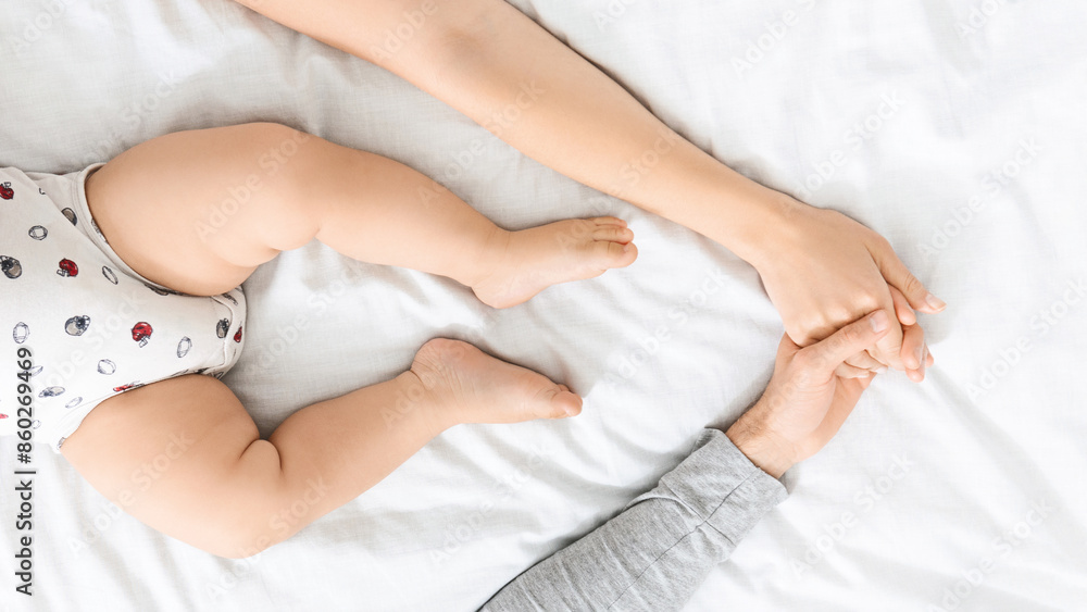 An overhead close-up photo of an adult and baby holding hands, lying on a white bed. The baby is wearing a white and red patterned onesie and is lying on its back with its legs in the air. An overhead close-up photo of an adult and baby holding hands, lying on a white bed. The baby is wearing a white and red patterned onesie and is lying on its back with its legs in the air.
