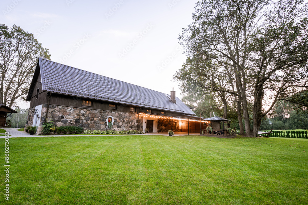 Blome, Latvia - September 11, 2023 - A scenic view of a rustic stone building with a modern roof, surrounded by lush green grass and trees, with outdoor seating and string lights. Blome, Latvia - September 11, 2023 - A scenic view of a rustic stone building with a modern roof, surrounded by lush green grass and trees, with outdoor seating and string lights.