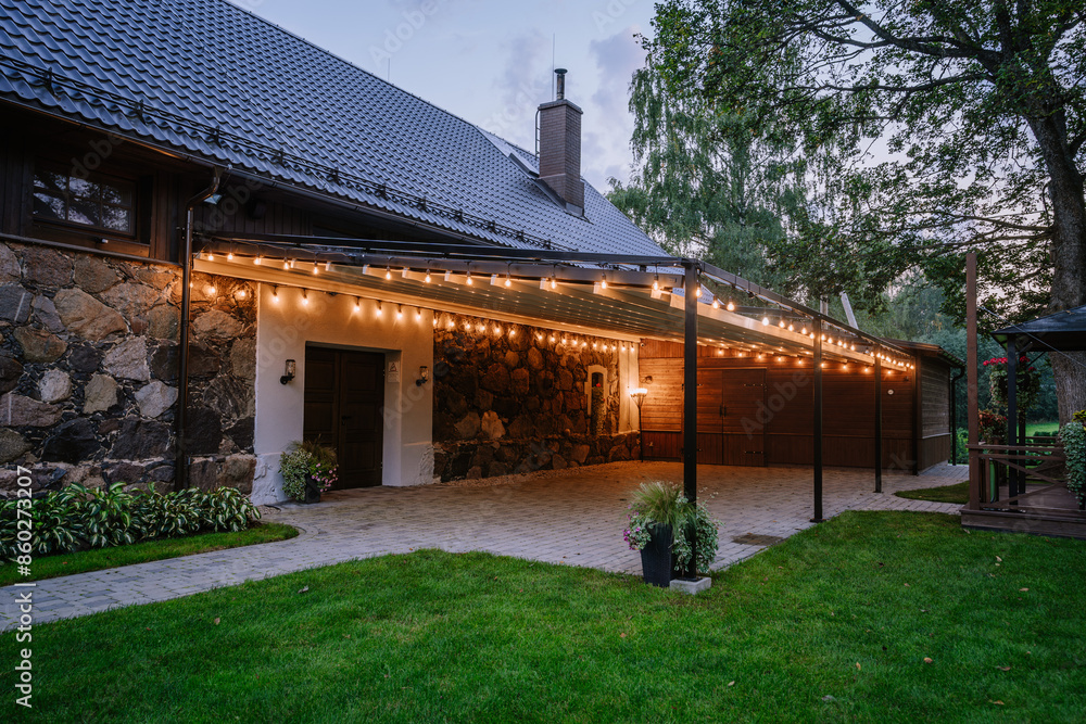 Blome, Latvia - September 11, 2023 - Outdoor covered patio area with string lights, attached to a stone and wooden building, surrounded by greenery and evening ambiance. Blome, Latvia - September 11, 2023 - Outdoor covered patio area with string lights, attached to a stone and wooden building, surrounded by greenery and evening ambiance.