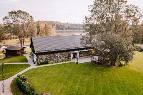 Blome, Latvia - September 11, 2023 - Aerial view of a charming stone house with a large green lawn, surrounding trees, and a cozy outdoor seating area, situated near a tranquil lake. photo