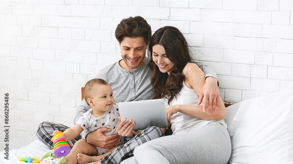 A happy family, including a young couple and their baby, is sitting on a bed in a bedroom with a white brick wall behind them. The couple is looking at a tablet, while the baby plays with a toy. A happy family, including a young couple and their baby, is sitting on a bed in a bedroom with a white brick wall behind them. The couple is looking at a tablet, while the baby plays with a toy.