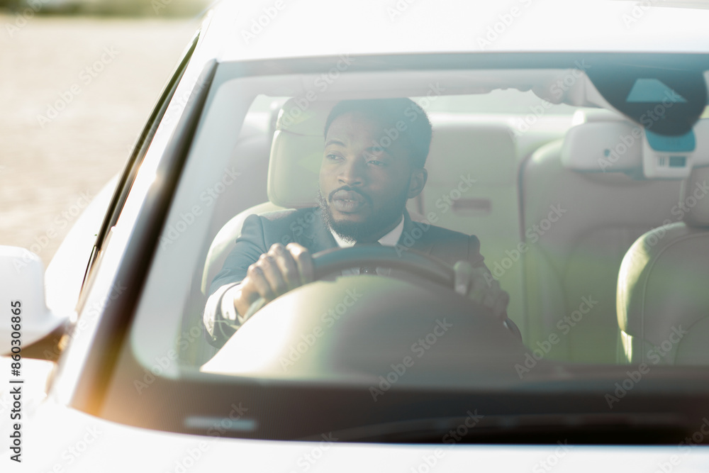 Naklejka premium Black man wearing a suit sits in the drivers seat of a car. The car is facing towards the sun, creating a bright, hazy effect. He is gripping the steering wheel and looking straight ahead