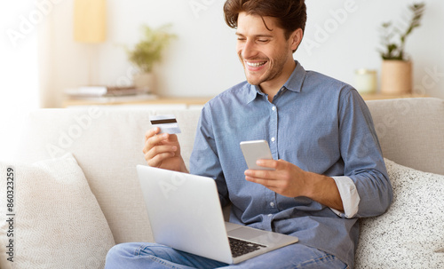 Obraz na płótnie A man on a couch smiles while looking at his phone holding a credit card, with a laptop in front, implying hes making an online payment