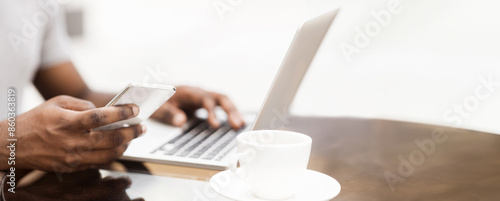 A close-up photo of a persons hands using a phone and a laptop. A coffee cup sits on a table to the right.