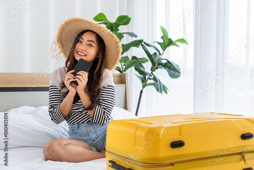 Young Asian woman wearing hat on vacation with smartphone and yellow suitcase in hotel room photo