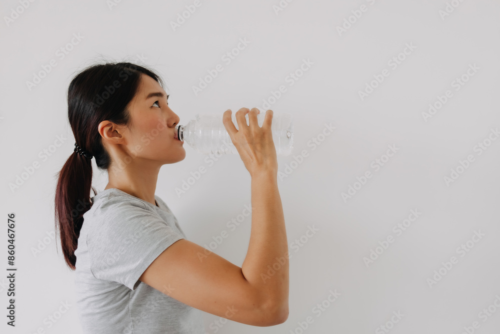 Asian Thai woman drinking mineral water from the bottle, standing isolated over white background wall. Asian Thai woman drinking mineral water from the bottle, standing isolated over white background wall.