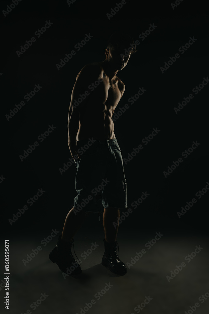 Muscular young man standing in a dark studio with dramatic lighting highlighting athletic physique. Male fitness, strength, and determination concept. Muscular young man standing in a dark studio with dramatic lighting highlighting athletic physique. Male fitness, strength, and determination concept.