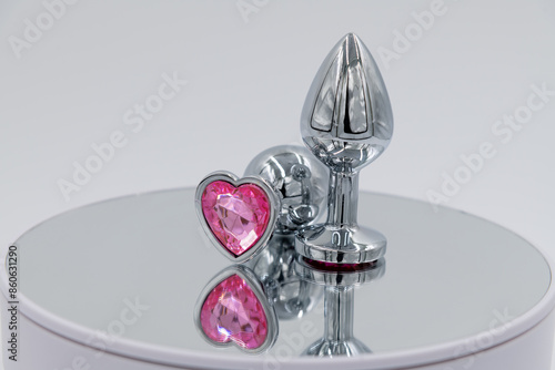 Close-up side view of two metallic butt plugs with pink heart shaped crystal lying on mirror surface against gray background. Soft focus. Adult sex toys theme. photo