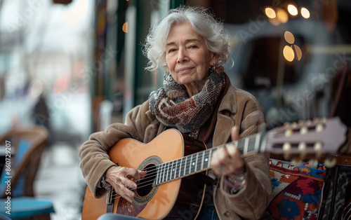 A senior woman sits outside and plays an acoustic guitar on a cloudy day. She wears a brown jacket and a scarf