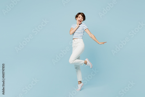 Full body sideways surprised fun young woman wears striped t-shirt casual clothes stand raise up leg look camera isolated on plain pastel light blue cyan background studio portrait. Lifestyle concept.