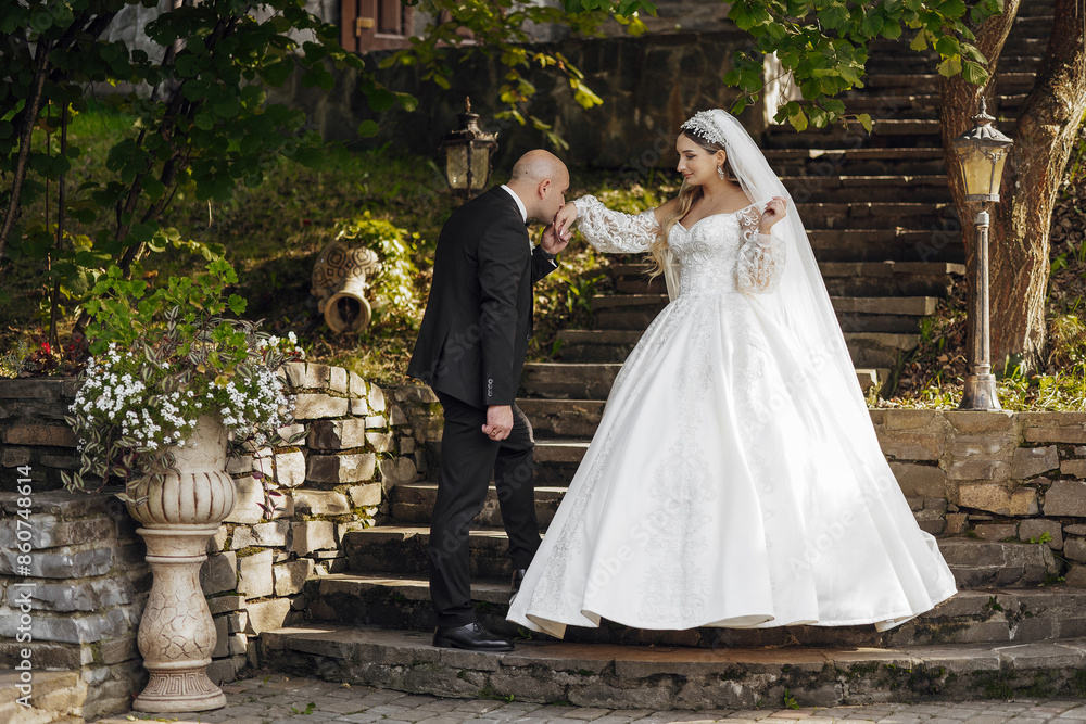 Fototapeta premium A bride and groom are walking down a stone staircase. The bride is wearing a white dress and the groom is wearing a suit. The scene is set in a garden with potted plants and a vase