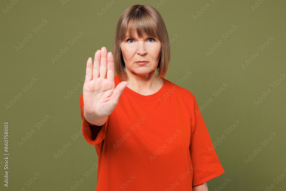 Elderly sad blonde woman 50s years old she wear orange t-shirt casual clothes look camera showing stop gesture with palm isolated on plain pastel green background studio portrait. Lifestyle concept. Elderly sad blonde woman 50s years old she wear orange t-shirt casual clothes look camera showing stop gesture with palm isolated on plain pastel green background studio portrait. Lifestyle concept.