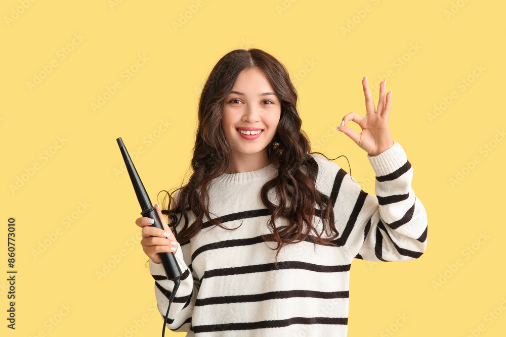 Young Asian woman with curling iron showing OK on yellow background Young Asian woman with curling iron showing OK on yellow background
