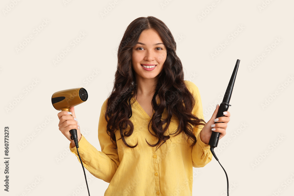 Young Asian woman with curling iron and hair dryer on light background Young Asian woman with curling iron and hair dryer on light background