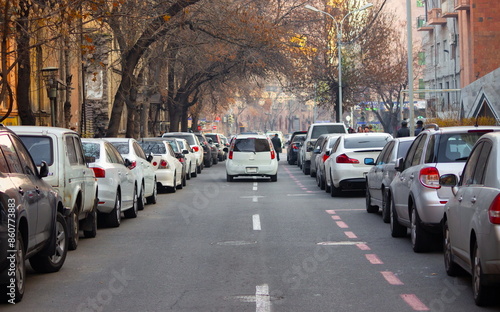 View of a city autumn street with parked cars on both sides, with buildings and trees in the backdrop