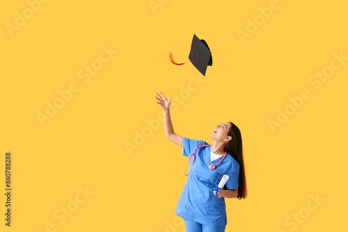 Happy female medical graduate with diploma and stethoscope on yellow background photo