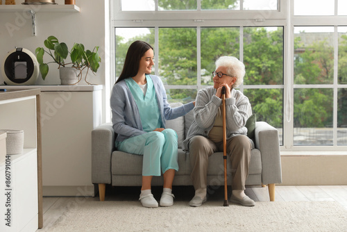 Young caregiver and senior woman with stick sitting on sofa in kitchen