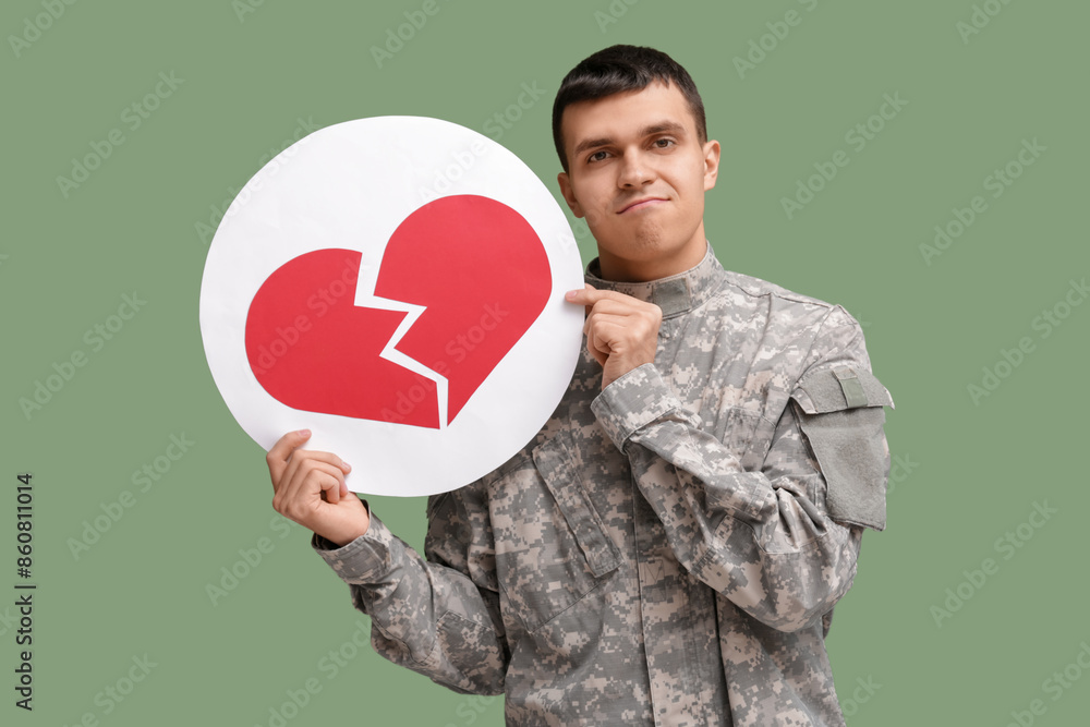 Handsome young displeased male soldier with heart dislike icon on green background