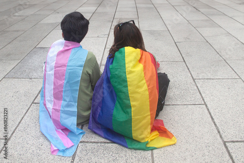 DOS CHICAS DE ESPALDAS, SENTADAS EN EL PISO DURANTE LA MARCHA DEL ORGULLO GAY, CON LAS BANDERAS COLGADAS SOBRE SU ESPALDA