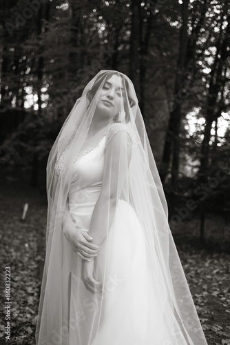 A woman in a white dress is standing in a forest, with a veil covering her face